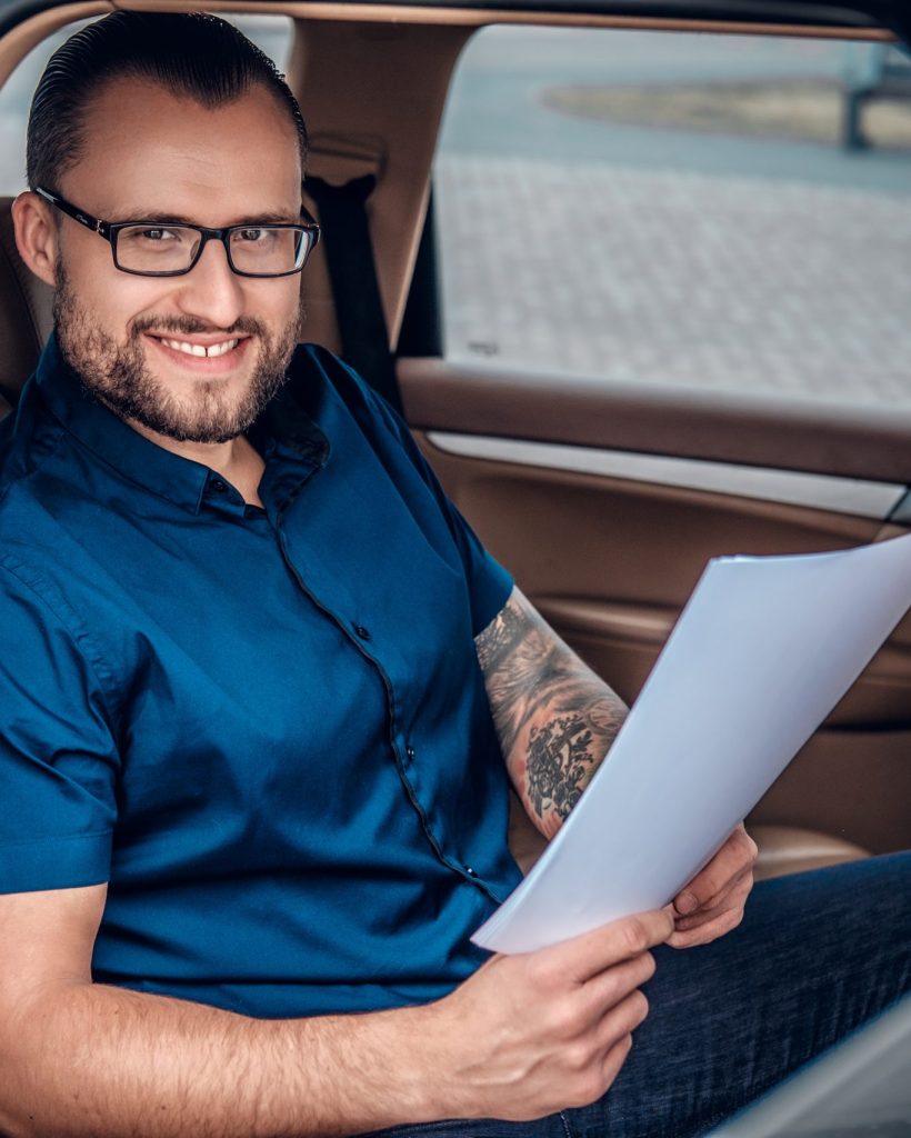 Bearded business male in eyeglasses with tattoo on his arm sits on a back seat of a car.